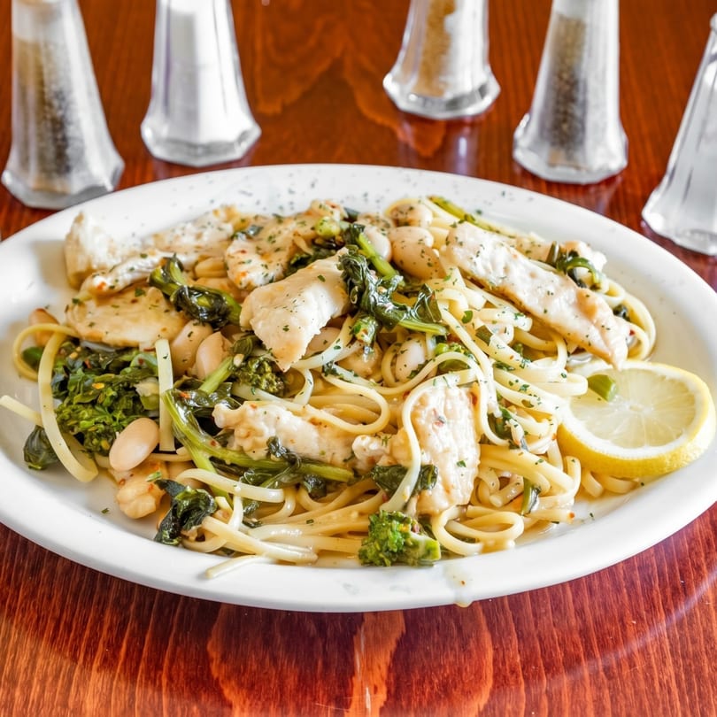 Chicken and broccoli rabe pasta with white beans and a lemon wedge on a white plate.