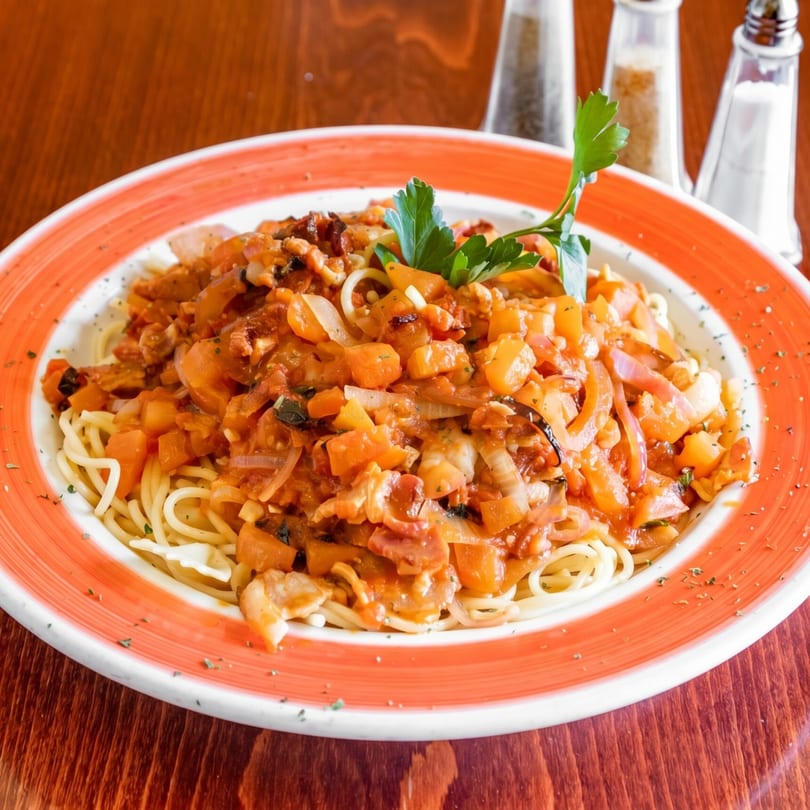 Spaghetti with chunky tomato sauce, onions, and bell peppers, garnished with parsley on a wooden table.
